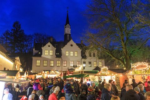 Weihnachtsmarkt mit vielen verzierten und geschmückten Ständen. Im Hintergrund ist ein großes weißes Gebäude zu sehen - gesäumt mit weißen Türmchen und Erkern.