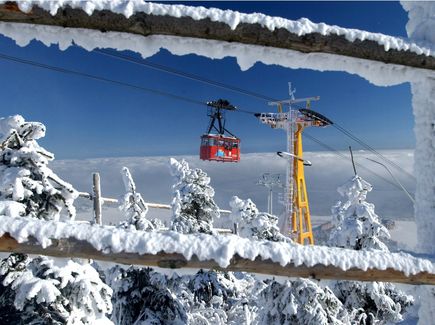 Blick durch einen verschneiten Zaun. Eine rote Schwebebahn fährt durch die Winterlandschaft des Erzgebirges. 