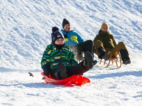 Familie rodelt im Winter einen Berg hinab. Es liegt Schnee und die Sonne scheint.