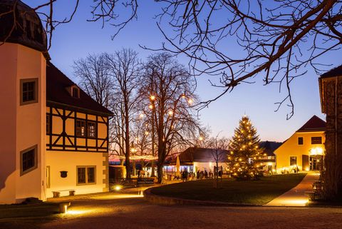 Das Bild zeigt einen Platz, auf dem dem in der Mitte ein Weihnachtsbaum steht. Ringsherum sind Häuser angeordnet. Lichterketten sorgen für eine anheimelnde Weihnachtsatmosphäre. Es ist abends, der Himmel ist verdunkelt.