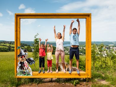 Eine Familie mit einem Mädchen und einem Jungen steht in einem goldenen Rahmen und lacht. Dahinter erstreckt sich die Landschaft von Eibenstock im Erzgebirge.