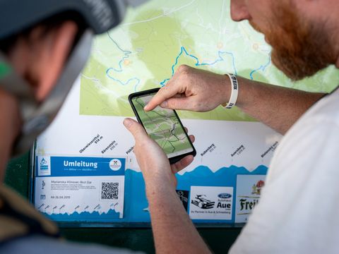 Zwei Mountainbiker stehen vor einer Übersichtskarte im Erzgebirge. Einer von ihnen hält ein Smartphone, auf dem ebenfalls die Karte zu sehen ist.