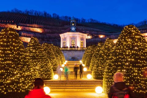 Ein weihnachtlich-festlich geschmückter Pavillon bildet das Zentrum des Bildes. Der Weg bergauf zum Pavillon ist gesäumt von Treppenstufen und geschmückten Bäumen rechts und links. Fünf Personen laufen die Treppen nach oben in Richtung Pavillon. 