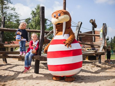 Eine Robbe mit einem rot-weiß gestreiften Anzug steht neben zwei Kindern auf dem Spielplatz. Sie sind im Ferienpark Trixi.