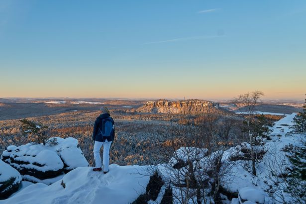 Eine Wanderin steht auf den schneebedeckten Felsen des Elbsandsteingebirges und blickt über die Winterlandschaft.