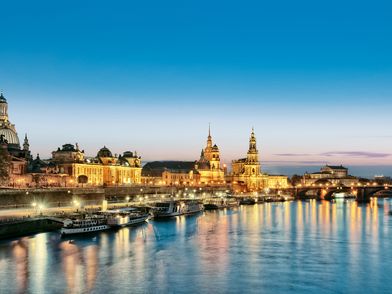 Am Ufer der Elbe befinden sich die prächtigen Gebäude der Dresdner Altstadt. Es ist Abend. Der Himmel färbt sich dunkel und überall leuchten Laternen.