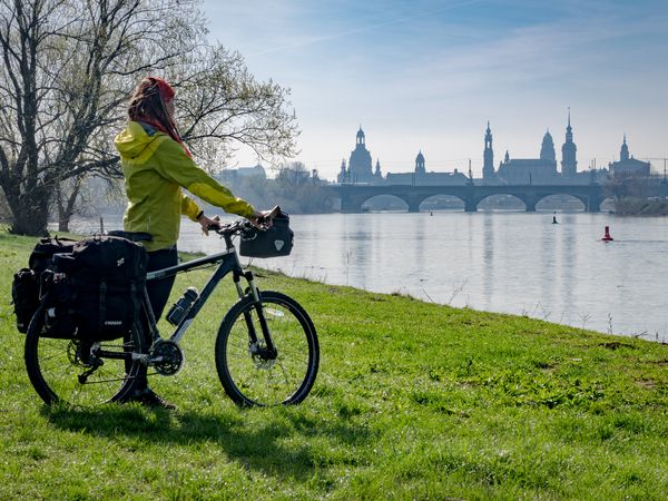 Eine Frau mit grüner Jacke steht neben einem Fahrrad. Sie schaut nach vorn auf den Fluss Elbe. Im Hintergrund befinden sich die berühmten Bauwerke der Stadt Dresden.
