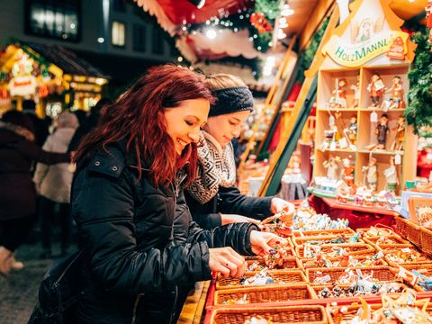 Zwei junge Frauen stehen vor einem Stand auf dem Leipziger Weihnachtsmarkt. Sie begutachten mit ihren Händen kleine Holzfiguren, die in Körben auf der Ladenfläche des Standes liegen. Der Stand ist weihnachtlich geschmückt und mit Lichtern umgeben. Im Hintergrund gehen weitere Besucherinnen und Besucher des Weihnachtsmarktes vorbei.