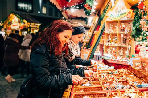 Zwei junge Frauen stehen vor einem Stand auf dem Leipziger Weihnachtsmarkt. Sie begutachten mit ihren Händen kleine Holzfiguren, die in Körben auf der Ladenfläche des Standes liegen. Der Stand ist weihnachtlich geschmückt und mit Lichtern umgeben. Im Hintergrund gehen weitere Besucherinnen und Besucher des Weihnachtsmarktes vorbei.