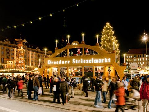 Ein Weihnachtsmarkt mit einem überlebensgroßen Schwibbogen im Zentrum mit dem Schriftzug "Dresdner Striezelmarkt" ist zu sehen. Es laufen zahlreiche Besucher und Besucherinnen über den Weihnachtsmarkt. Die Schaubuden und Stände leuchten durch angebrachten Weihnachtsschmuck.