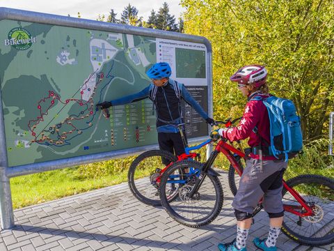 Ein Mann und eine Frau stehen mit ihren Mountainbikes an einer Tafel in der Bikewelt Schöneck im Vogtland.