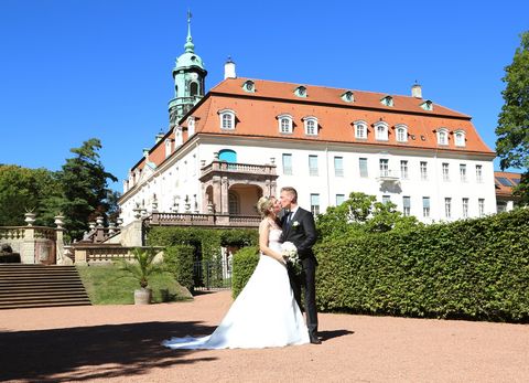 Ein Mann in einem schwarzen Anzug küsst eine Frau in einem weißen Brautkleid. Im Hintergrund befindet sich eine Hecke und das Schloss Lichtenwalde. 