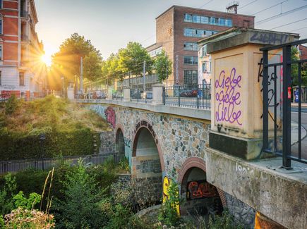Eine Steinbrücke führt über Wasser. Es ist Abendstimmung. Die Sonne über der Stadt geht langsam unter. Die Brücke ist mit Graffiti bemalt.