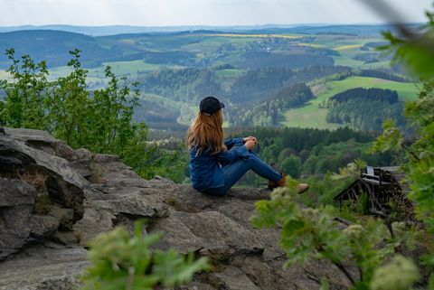 Eine Frau sitzt auf einem Felsen und schaut in die Natur des Erzgebirges. Sie ist an einer Aussicht des Erzgebirger Kammwegs.