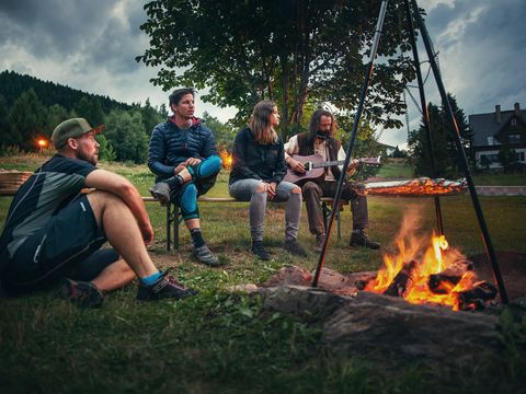 Eine Gruppe sitzt gemütlich am Lagerfeuer zusammen und ein Mann spielt ein Lied auf der Gitarre. 